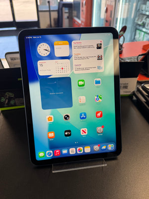 An Apple iPad tablet on a clear display stand, showing the home screen with various apps and widgets. The tablet is photographed on a dark tabletop in a retail or office environment with blurred background elements.