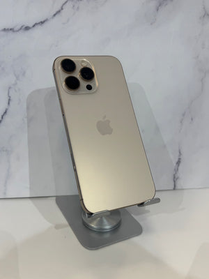 An Apple iPhone 12 Pro in light gold color, standing upright on a gray metal display stand against a white marble-patterned background.