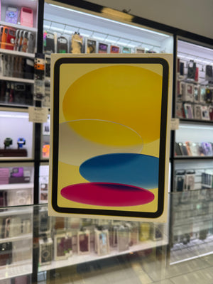 A tablet box standing upright on a glass display counter in a retail store. The front features an abstract design with large overlapping yellow, blue, and pink shapes inside a black border, with shelves of electronics in the blurred background.