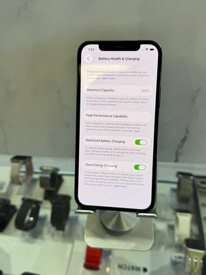 A black smartphone displayed on a metal stand, showing the 'Battery Health & Charging' settings screen on its home screen. The phone is set against a blurred retail background with a marble-patterned wall.