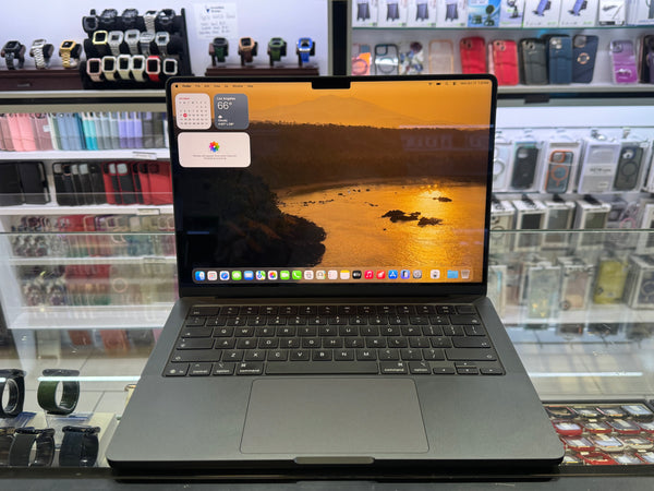 A dark gray Apple MacBook laptop is open and centered on a retail display counter, with its screen on showing a macOS desktop and a weather widget. The background shows shelves of various smartwatches and phone cases in a store.