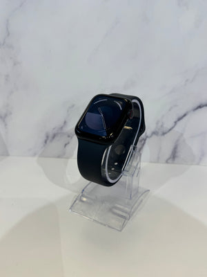 A black smartwatch with a rectangular digital display and black band, displayed on a clear acrylic stand against a white marble-patterned background.