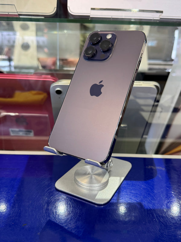 A purple iPhone 12 Pro Max with triple rear cameras, resting on a silver metal display stand on a glossy blue counter inside a retail store.