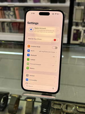 A black smartphone displaying the Settings app screen, photographed in a retail or showroom environment with glass surfaces and blurred electronics in the background.