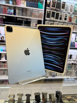 An Apple iPad Pro shown in a retail store setting, with the back of a gold-colored iPad standing upright on a clear display stand and a second iPad leaning against it showing its colorful screen.