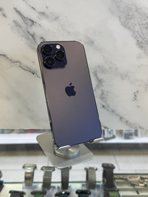 A dark purple Apple iPhone 12 Pro displayed on a silver stand, showing its back with the triple-camera array and Apple logo, set against a white marble-patterned background.