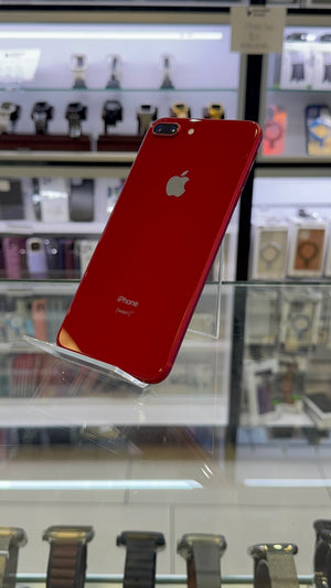 A red Apple iPhone 7 Plus displayed on a clear stand inside a retail store, showing the back of the phone with the Apple logo and 'iPhone' text.