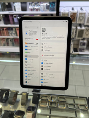 A black tablet displaying the Apple Settings app, specifically the Apple Account General page, resting on a glass display counter in a retail store.