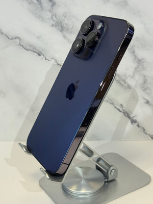 A dark blue iPhone 12 Pro Max displayed on a silver metal stand, showing the back of the phone with its triple rear cameras and Apple logo, set against a white marble-patterned background.
