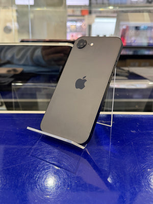 A dark gray Apple iPhone shown from the back, mounted on a clear display stand against a bright blue wall in a retail or showroom setting.
