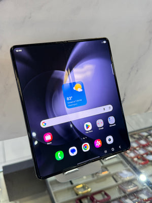 A black foldable smartphone displayed on a stand, showing its screen on in a retail setting with a marble wall and glass display case in the background.