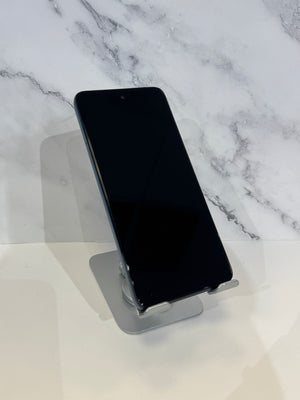 A black smartphone with a front-facing camera cutout stands upright on a clear acrylic display stand against a white marble-patterned background.