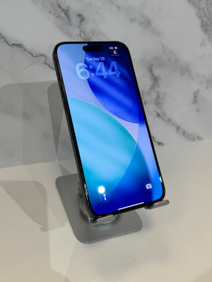 A black iPhone with a blue and teal lock screen wallpaper displayed on a clear acrylic stand with a gray base, set against a white marble-patterned background.