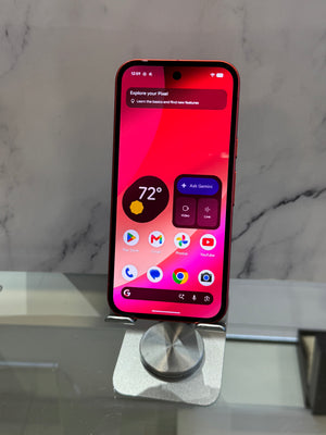 A smartphone with a black front and red side edges stands upright on a small silver metal display stand. The screen is on and shows a colorful home screen with weather and app icons.