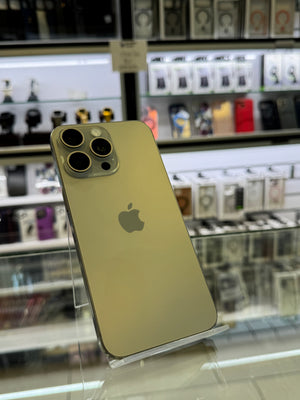 A gold-colored Apple iPhone Pro displayed on a clear stand inside a retail store, with blurred shelves of other phones in the background.