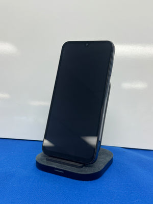 A black smartphone with a blank screen stands upright on a dark gray charging dock against a white background, with a blue surface beneath the dock.