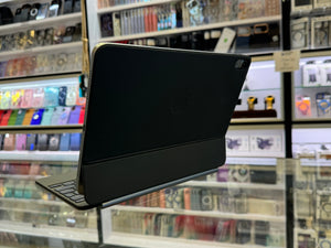 A black iPad in a protective folio case with an attached keyboard, shown from the back inside what appears to be a retail store with shelves of phone accessories in the background.