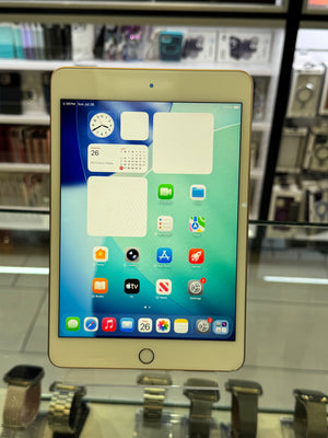 A light gold Apple iPad shown from the front with its screen turned on, displaying the home screen with various app icons. The device is photographed inside what appears to be a retail display or showroom with glass cases and metallic items in the background.