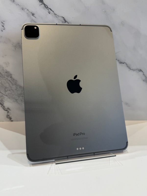 The back of an Apple iPad Pro in a dark metallic gray finish, resting on a clear acrylic stand against a white marble-patterned background.