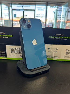 A blue Apple iPhone with a black protective case stands on a dark charging dock on a black table. Behind it are two black and green ESOUK electric car charger boxes, with a blurred storefront background.