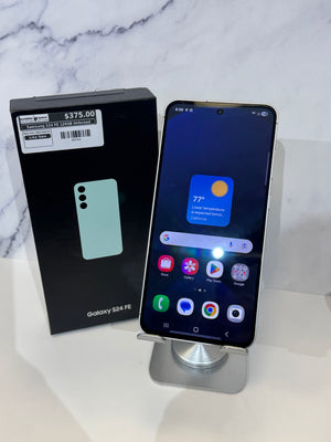 Samsung Galaxy S24 FE smartphone displayed on a silver stand next to its original black retail box on a white surface with a marble-patterned background.
