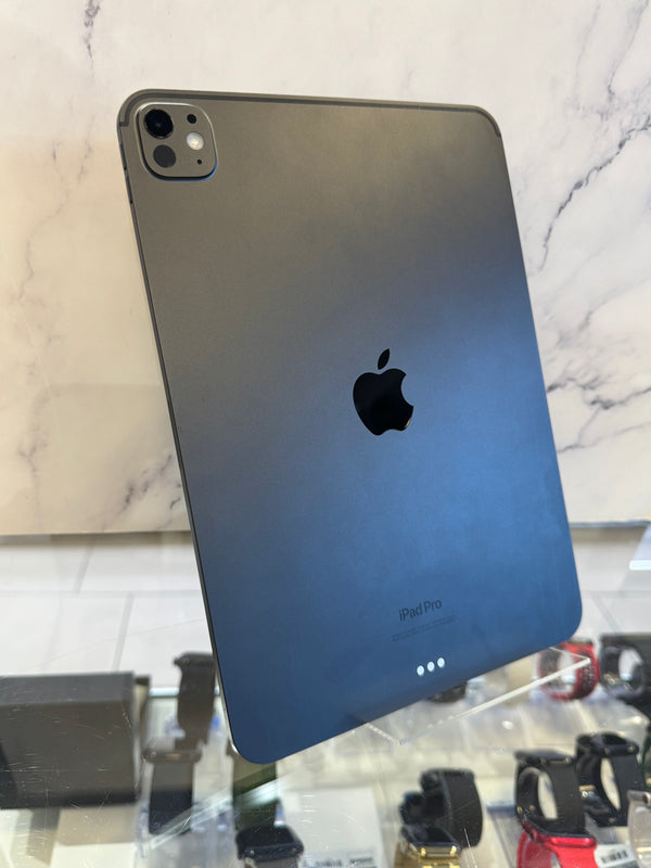 A dark gray Apple iPad Pro shown from the back, standing on a clear display stand in a retail or showroom setting with marble wall panels and other accessories visible in the background.