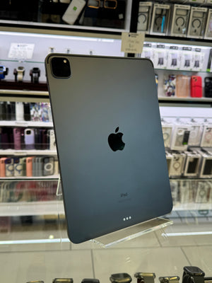A dark gray Apple iPad shown from the back, resting on a clear display stand inside a retail store with shelves of accessories visible in the background.