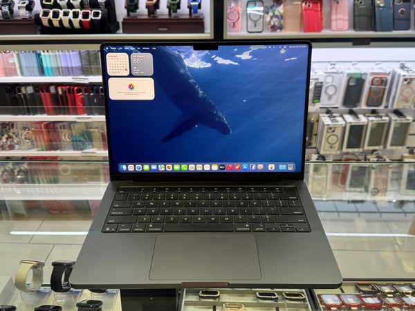A dark gray Apple MacBook laptop with an open screen displaying a whale wallpaper, set on a glass display counter inside a retail store.