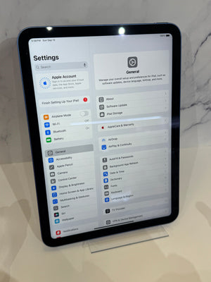 A black tablet, likely an iPad, standing upright on a clear display stand on a light marble surface. The screen is on and displaying the Apple Settings interface, with the General settings page visible.
