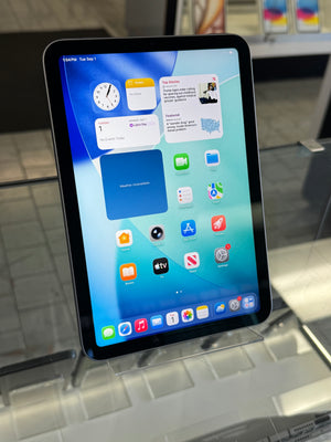 A black tablet with a blue and white home screen displayed on a clear stand in a store setting.
