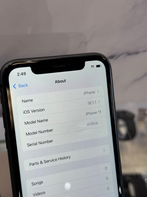 A close-up of a black smartphone displaying the 'About' screen with iOS version and device details, set against a blurred light gray marble background.