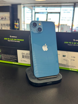 A blue Apple iPhone shown from the back, resting on a dark charging dock or stand. The Apple logo is visible in the center, and the phone is positioned in front of its retail packaging box on a dark tabletop.