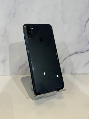 A black Samsung Galaxy smartphone shown from the back, resting on a clear acrylic display stand against a white marble-patterned background.