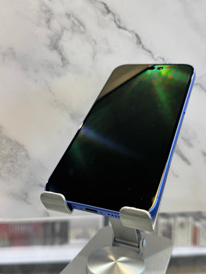 A blue smartphone with a glossy black screen resting on a silver metal stand, set against a white marble-patterned background.