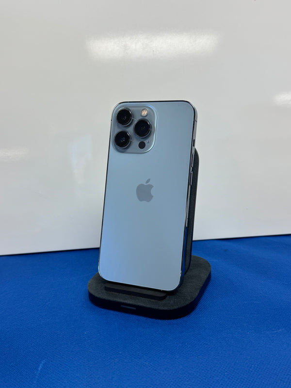 A blue Apple iPhone Pro standing upright on a black charging stand against a white wall, with a blue surface below.
