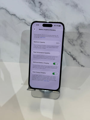 A black smartphone displayed on a silver metal stand, showing the battery health and charging settings screen. The phone is set against a white marble-patterned background.