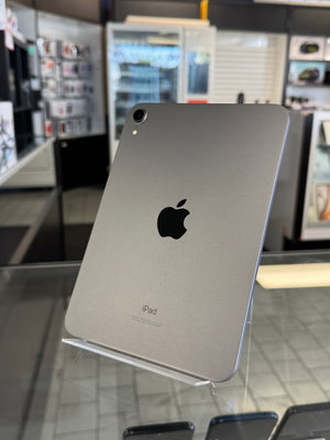 A silver Apple iPad shown from the back, resting on a clear display stand inside a retail store. The Apple logo is clearly visible in the center.