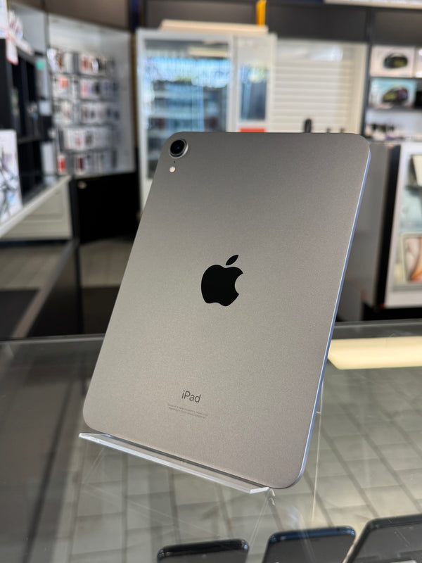 A silver Apple iPad shown from the back, resting on a clear display stand inside a retail store. The Apple logo is clearly visible in the center.