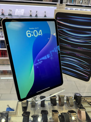 A tablet device with a black bezel and a blue-to-green lock screen wallpaper, photographed in a retail display area with watches and another device visible in the background.