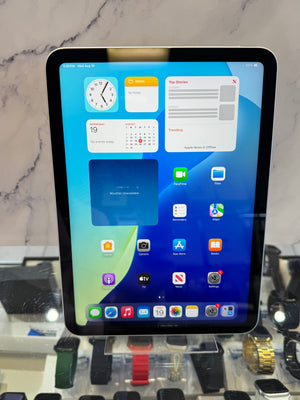 A black Apple iPad with a large screen displaying various colorful app icons and widgets, standing on a clear display stand in front of a marble-patterned wall.