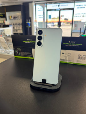 A light green GeekGeek smartphone displayed on a black charging stand on a dark counter inside a retail store. GeekGeek branded product boxes are visible in the background.