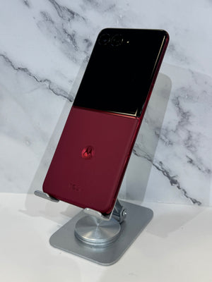 A Motorola flip-style smartphone with a glossy black upper half and deep red lower half, displayed on a silver stand against a white marble-patterned background.