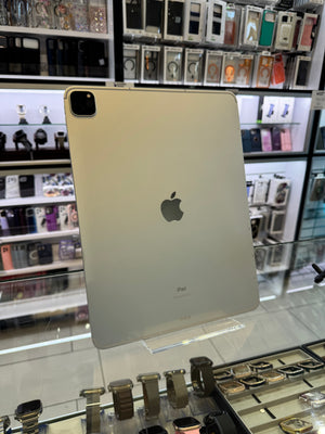 A silver Apple iPad Pro shown from the back, standing on a clear display stand inside a retail store. The Apple logo is visible in the center, with shelves of phone cases and other electronics in the background.