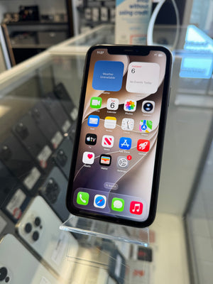 A black iPhone with a lit home screen displaying various app icons, resting on a clear acrylic display stand inside what appears to be a retail store.