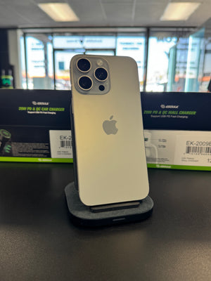 A silver Apple iPhone stands upright on a small black charging stand on a dark counter, with Apple charger boxes visible in the blurred background.
