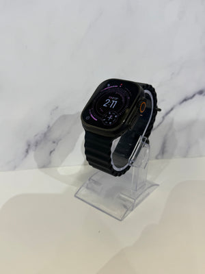 A black smartwatch with a rounded rectangular face and black strap, displayed on a clear acrylic stand against a white marble-patterned background.