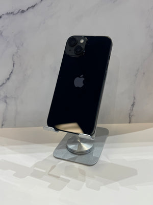 A black Apple iPhone with a glossy finish is displayed on a silver metal stand, set against a white marble-patterned background.