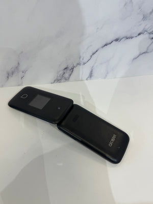 A black flip phone with a small screen on the outer cover and a visible Alcatel logo on the inner cover, resting on a white surface against a white marble-patterned background.