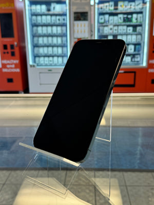 A black smartphone with a blank screen resting on a clear acrylic display stand in a retail or showroom setting, with vending machines and tiled flooring blurred in the background.