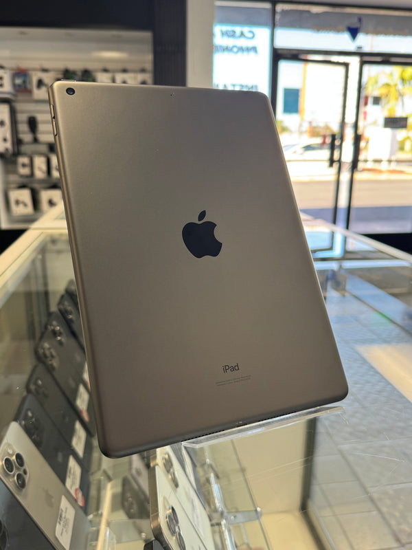 A silver Apple iPad shown from the back, resting on a glass display counter inside a retail store. The Apple logo is clearly visible in the center.